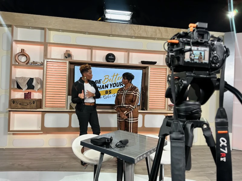 behind the scenes of a TV studio with the camera in view and two women standing in the distance on stage in front of a wall with a TV and shelves.